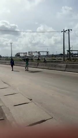 The video shows a wide, grey, concrete road with concrete barriers in the middle. The sky is partly cloudy. In the distance, there are constructions, electrical posts, and what appear to be industrial areas.

In the beginning, two men are walking along the side of the road. One man carries a large, flat object above his head. A motorcycle passes them. The camera then pans to the side of the road, where more motorcycles are parked, and people are standing around. 

The camera continues to pan, showing a busy area with yellow vans, three-wheeled vehicles, and people. There are some shops with signs.