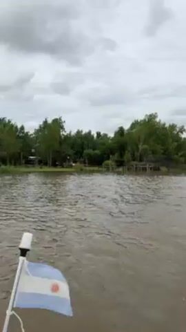 The video shows a boat trip on a body of water near a shoreline under an overcast sky. The Argentinian flag is seen flapping on the front of the boat. The water is dark and choppy. The shoreline is densely covered in trees and vegetation. A few small, simple dwellings are visible along the shore, some slightly elevated on stilts, suggesting they are designed to cope with potential flooding. One person is visible in the foreground, but their activity is unclear. The overall impression is a somewhat remote, possibly rural or delta region.