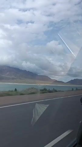 The video shows a view from inside a moving vehicle, looking out the side window. The scene outside consists of a highway with a concrete barrier, a body of water, and mountains in the distance under a cloudy sky. There are reflections on the car window, including what appears to be the interior of the car and a white object.