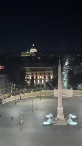 The video shows a night-time aerial view of a large, open square in a city. A prominent obelisk stands in the center of the square, illuminated by lights. Several people are visible walking around the square and gathered near fountains around the obelisk. In the background, buildings are lit up, and a dome-shaped structure, possibly a church or basilica, is visible in the distance. The sky is dark, suggesting it is nighttime. The overall atmosphere is peaceful and illuminated.