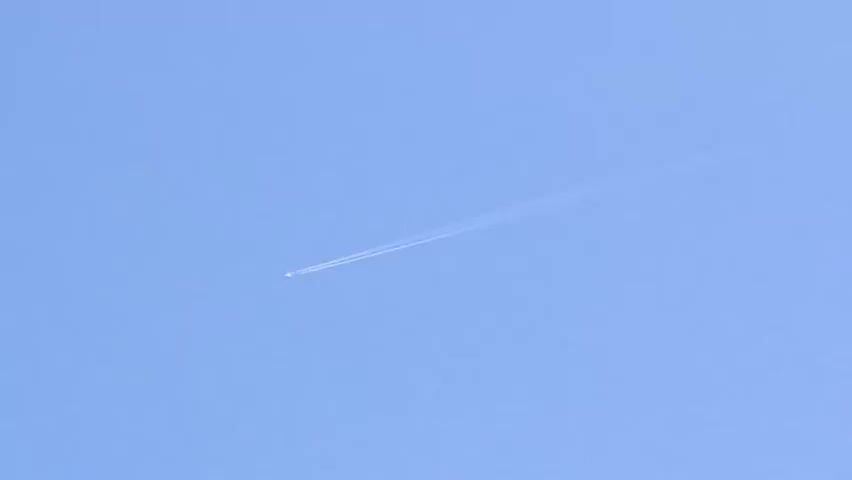 The video shows a clear blue sky with a single airplane flying diagonally across the frame, leaving a white contrail behind it. The plane appears small and distant, and the contrail is thin and wispy, suggesting it is at a high altitude. The sun is not visible, but the sky is brightly lit, indicating daytime. There are no other objects or people visible in the frame. The camera remains stationary throughout the recording.