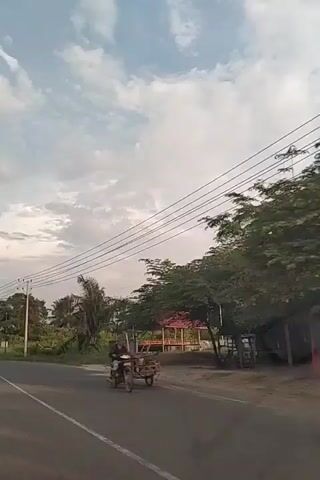 The video captures a slightly tilted, side-view perspective of a road, likely in a tropical or subtropical region, as indicated by the lush vegetation and palm trees. The sky is partly cloudy with a mix of white and gray clouds, suggesting it might be late afternoon or early evening. Power lines stretch across the frame, supported by poles, and a few residential structures are visible along the roadside, including one with a red roof. In the foreground, a person on a motorcycle is blurred due to motion, moving from right to left across the frame. The road surface appears to be asphalt, with a white line marking the edge of the lane. The overall impression is that of a rural or semi-rural setting with active, but calm, local life.