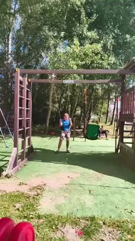 A boy is swinging on a swing set outdoors. He is wearing a blue shirt and patterned shorts. He is in the center of the frame, swinging back and forth. The swing set is made of wood and has a ladder on the left side. There is green artificial turf on the ground. In the background, there are trees, other playground equipment, and another person sitting down. The sky is visible and appears to be clear.