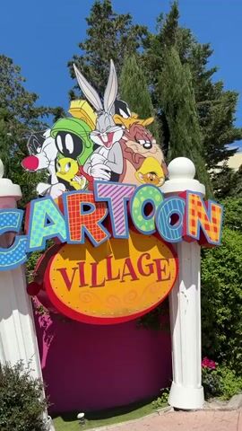 The video shows a "Cartoon Village" sign featuring Looney Tunes characters.  A pink wall is seen in the background. People walk around a paved area lined with trees.  A roller coaster is visible in the background. A statue of the Tasmanian Devil is also shown.