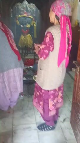 The video shows two women standing inside a dimly lit room, possibly a temple. The woman on the right, wearing a pink headscarf and a light-colored vest over a floral print outfit, is holding a lit diya (oil lamp) and appears to be offering prayers to a dark statue in an alcove. The statue is adorned with gold fabric and garlands of flowers. The woman on the left, facing away from the camera and also wearing a headscarf and a grey overcoat over a pink salwar kameez, stands slightly behind the statue. The floor is tiled, and there are offerings and other items on the ground in front of the statue. The overall atmosphere is one of religious devotion.