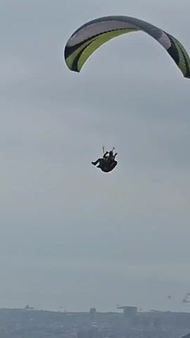 A paraglider floats in the sky with a grey and green canopy, suspended above a city obscured by haze and a sand dune. The individual is centered in the frame, their form silhouetted against the overcast sky. The horizon shows a dense urban sprawl fading into the distance, and the sand dune is in the foreground. The overall tone is muted, with a flat, cloud-covered sky.