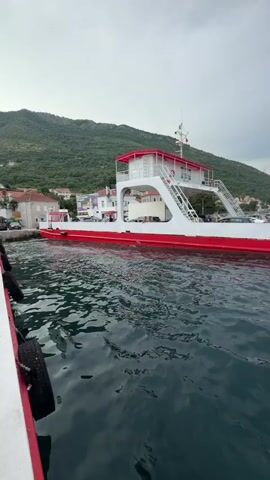 A ferry boat is docked in the water, with a town and green hills in the background. The ferry is white with red accents. There are cars parked on the deck of the ferry. The water is calm, and the sky is overcast.