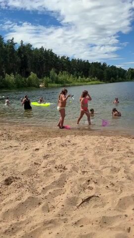 This media file captures a group of children enjoying a summer day at a beach near Stūnīši, Latvia. The scene is set on a sandy shore bordering a calm lake, with lush green trees forming a backdrop and a bright blue sky with scattered clouds overhead.

The primary subjects are several children, all wearing swimwear, who are actively engaged in water play. In the foreground, a young girl in a light blue and pink swimsuit is running towards the water, her sandy feet kicking up dust. As the camera pans, we see other children entering the lake. One girl in a purple swimsuit is splashing energetically, holding a pink net. Another girl, in a pink bikini, is already in the water, appearing to be playing with the others. Several other children are visible in the lake, some swimming and others wading.

The activity depicted is clearly recreational, focusing on the children's enjoyment of a sunny day at the beach. The overall mood appears to be one of joy and carefree fun, amplified by the energetic splashes and the children's evident excitement. The time of day is likely midday or early afternoon, given the bright sunlight and clear sky. The location cues, such as the sandy beach, the lake, and the surrounding forest, strongly suggest a natural outdoor setting suitable for swimming and beach activities. There is no visible text in the media file.