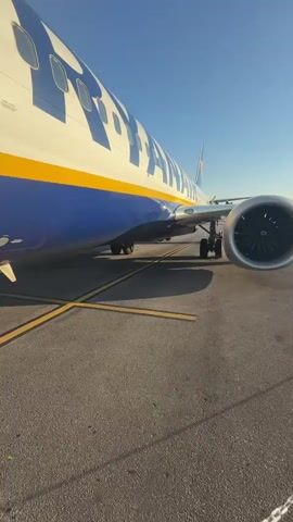 The video shows a close-up, ground-level view of the side of a Ryanair airplane parked on an asphalt tarmac. The plane's white fuselage is marked with large blue and yellow Ryanair lettering. A blue and yellow stripe runs horizontally along the bottom of the fuselage. The airplane's landing gear is visible, as is one of its jet engines, which is silver with black fan blades. Yellow lines on the tarmac are visible in the foreground. The sky above is clear and blue. The camera is positioned low, near the ground, looking up at the airplane.