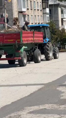 A blue tractor pulls a green and red trailer loaded with debris down a street. Two individuals are seated in the tractor's cab. Another two individuals are visible walking in the background on the right side of the frame. The scene takes place in an urban environment with multi-story buildings lining the street. The road surface appears to be asphalt or similar paving material, with distinct markings visible. The weather appears to be sunny.