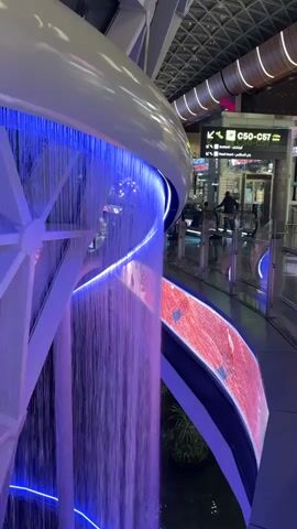 The video shows a modern, indoor waterfall feature with purple and blue lighting. The waterfall cascades down a curved structure. In the background, an airport or large public building is visible, with signage indicating gates C50-C57. Several people are walking in the background, some near security gates. A person is standing near a gate, and another person is walking past. The environment appears to be well-lit and spacious.