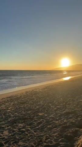 A beach at sunset. The sun is setting over the ocean, casting a warm glow on the sand and water. Waves are gently rolling onto the shore. The sky is clear and blue with a hint of orange and yellow from the setting sun. There are no people or objects visible in the video, only the natural beauty of the beach and the ocean.