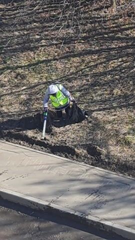 From an elevated angle, the video captures a person picking up litter. The individual is clad in a gray hoodie, a high-visibility yellow vest, dark pants, and dark shoes. They're employing a litter picker to grasp refuse and deposit it into a black trash bag. The surroundings comprise a concrete sidewalk and a dirt and grass area dotted with branches and shadows from a tree or shrubs.