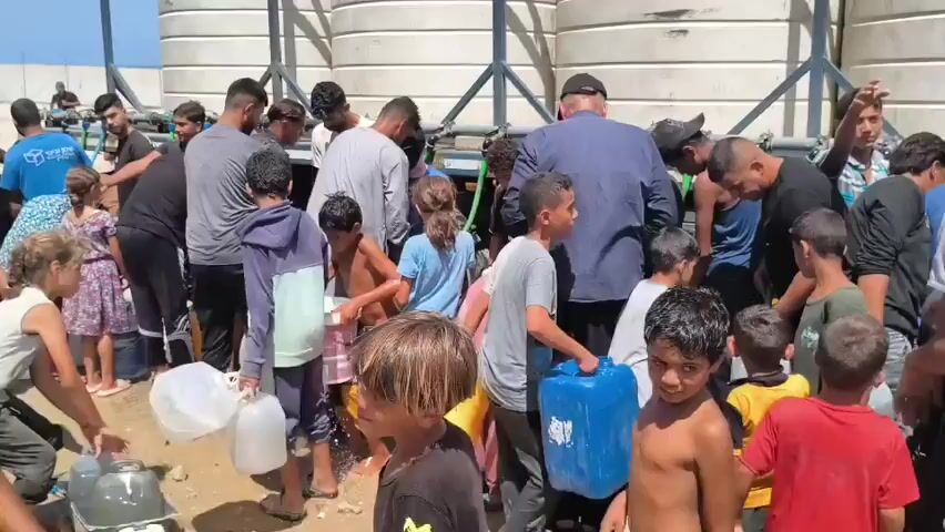 A group of people, including many children and some adults, are gathered outdoors near large white cylindrical tanks. They are collecting water from hoses and pipes connected to the tanks into various containers, including large plastic jugs and buckets. The ground is dirt and sand, and there is some vegetation and fencing visible in the background. The atmosphere appears to be one of necessity and communal effort, possibly indicating a water shortage or distribution point. People are actively engaged in filling their containers and assisting each other.
