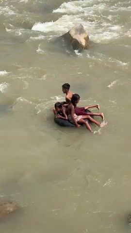 Three children are playing in a fast-moving river, seated in an inflatable inner tube. The water is murky brown, and there are rocks visible both in and out of the water. The children appear to be enjoying themselves as they navigate the currents. One child is at the front, facing forward. Two other children are seated behind, one facing forward and the other facing backward. Their legs are dangling in the water. The scene is captured from an elevated angle.