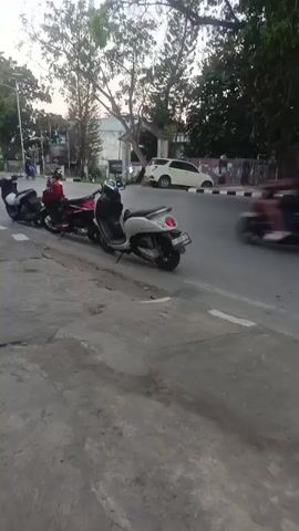 The video shows a street scene with several motorbikes parked along the side of the road. A white car is visible in the background, driving on the road. The sky is overcast, and there are trees and buildings along the street. The camera pans slowly to the left, showing more of the parked motorbikes and the surrounding environment. The overall scene appears to be a typical urban street during daylight hours.