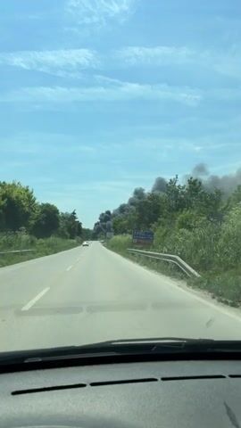 A video of a road with a large cloud of black smoke on the right side of the road. There are several cars on the road. The sky is blue with a few clouds. The video is taken from inside a car.