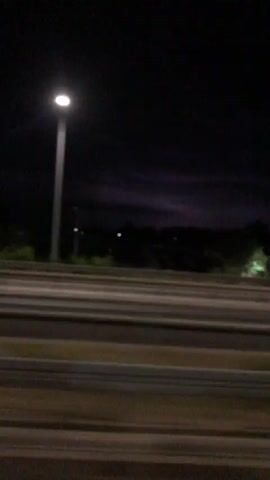 A first-person perspective shot from inside a vehicle at night. The camera is looking forward and slightly to the right. In the foreground, there are three lanes of a road, separated by what appear to be curbs or dividers. Beyond the road, there is a grassy median with several tall streetlights, some of which are illuminated. In the distance, there are faint lights that suggest buildings or structures. The sky is completely dark, indicating nighttime. The video is somewhat blurry, suggesting motion.