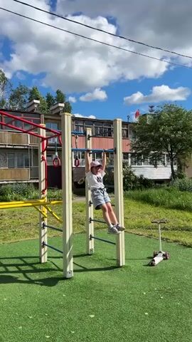 The video shows a young girl on a playground. The girl, wearing a white t-shirt, denim shorts, and a light pink baseball cap, is hanging from a set of horizontal bars on a play structure. She is moving her hands along the bars, traversing the structure while suspended. To the side of the structure is a scooter parked on the green artificial turf of the playground surface. In the background, there are apartment buildings, trees, and a blue sky with scattered clouds. A red flag flutters atop one of the buildings. The girl calls out to someone, saying "Mom, Mom!"