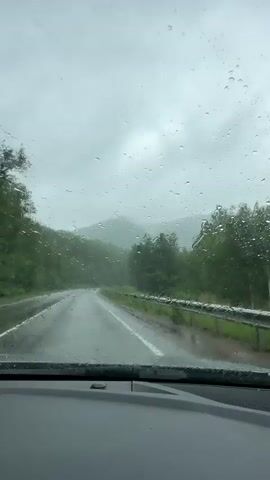 The video shows a view from inside a car driving on a wet road through a mountainous, forested area. Raindrops are visible on the windshield, distorting the view of the road ahead and the surrounding trees and hills. The road curves to the left, and a guardrail is visible along the right side of the road. The sky is overcast and appears to be gray. The lighting is dim, consistent with a rainy day. No people or other vehicles are visible.