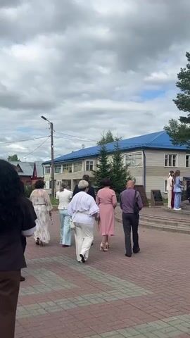 The video shows a group of people, likely students, dressed in formal attire including suits and dresses, walking up a set of stairs towards a large, modern, golden structure. The architecture of the building in the background, with its blue roof and light-colored siding, suggests a civic or institutional building, possibly a school or administrative office. The weather appears to be overcast, with a cloudy sky, and the time of day seems to be daytime. The atmosphere appears to be celebratory or formal, given the dress code and the organized gathering. Some individuals are holding flowers, which could indicate a special occasion like a graduation or a ceremony. There are also adults present, some appearing to be teachers or chaperones. A man in a uniform, possibly a military or ceremonial guard, stands near the golden structure, adding to the formality of the event. The paved walkway and stairs suggest an outdoor public space. The overall impression is of a significant event being held outdoors in a community setting.