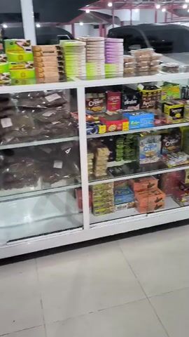 The video shows the interior of a small shop or store that sells snacks and sweets. The camera pans across display cases filled with various packaged goods. In the first display case, from left to right, are stacks of colorful boxes, then rows of packaged snacks like cookies and crackers. The second display case, to the right, is also filled with packaged snacks, with some shelves appearing empty. Towards the end of the video, a person wearing a hijab and a patterned top is visible, reaching into one of the display cases. The environment appears to be an indoor retail space with tiled flooring and fluorescent lighting.
