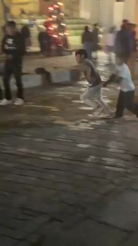 The video shows an outdoor scene at night, likely in a public square or plaza, where several children are playing soccer with a green ball on a paved surface. There is a lit-up Christmas tree in the background, decorated with ornaments. Other people, including adults and children, are visible in the background, sitting or walking. A stroller is parked nearby. The area seems to be illuminated by streetlights.