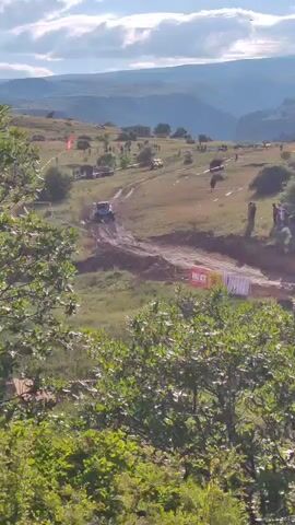 A four-wheeled vehicle is seen driving on a dirt track with spectators on the side of the track. The vehicle is kicking up dust as it drives. The scene appears to be a race or competition. The terrain is hilly and there are trees in the foreground. The sky is cloudy.