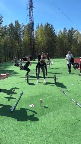 The video captures a group workout session on a bright, sunny day in what appears to be an outdoor training facility with artificial turf. A woman in black and patterned leggings is at the center of the frame, performing a weightlifting exercise with a barbell, lifting the weight overhead. Around her, other participants are engaged in various activities including squats with barbells, seated exercises, and walking. Several other barbells are lying on the green turf. The area is surrounded by trees and a fence, and a tall metal structure, resembling a communication tower, stands in the background. A red object, possibly a car part or equipment, is visible to the right side of the frame. Someone in a white shirt walks in front of the camera. The overall atmosphere is active and fitness-oriented.