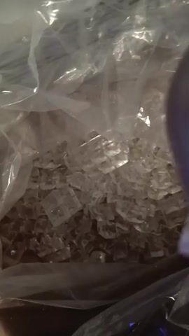 The video shows a clear plastic bag filled with ice cubes. The bag is opaque in some areas and translucent in others, allowing the ice to be seen within. The ice cubes appear to be small and uniformly shaped, possibly from an ice maker. The bag is placed on a surface that appears to be a wooden countertop. In the background, there is a stainless steel appliance with vents, possibly a refrigerator or an ice machine. The lighting in the scene is dim, but the ice cubes reflect the light, making them appear bright and shiny. The camera is focused on the bag of ice, and the movement of the camera is minimal. There are no people or other objects visible in the video.