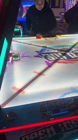 In an arcade setting, two people are actively engaged in a game of air hockey. The air hockey table, brightly lit and featuring colorful graphics with text like "OPEN ICE" and "QUAD-AIR," is the central focus. One person, primarily seen from the back, is on the left side of the table, holding a red air hockey mallet. On the right side, another person is also playing, using a red mallet to strike a puck. Several pucks are visible on the table, including a green one near the center and a yellow one on the left. A man in a black hoodie stands behind the player on the right, watching the game with a smile on his face, indicating enjoyment. Another woman is partially visible behind the man, also observing the game. The background is filled with various arcade machines, displaying bright, colorful lights, consistent with an entertainment venue. A small digital display showing "02" is visible near the left side of the table.