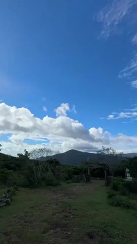 The camera pans across a scene of a lush green landscape under a bright blue sky with scattered white clouds. The foreground consists of a grassy field dotted with trees and shrubs. In the middle ground, there's a glimpse of what appears to be a simple wooden structure. Behind the trees, a mountain range is visible, partially obscured by the clouds.