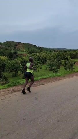 A bald Black man runs on a paved road away from the camera. He wears a light green sleeveless shirt with the number 13 on the back and dark running shorts. He runs with a good rhythm and posture. The road has cracks in it. The man runs past a line of shrubs and green grass that separates the road from a field of lush greenery. Beyond the field is a green hill against a backdrop of a cloudy sky.