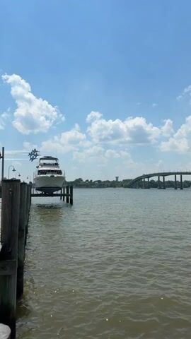 The video shows a body of water with a bridge in the distance. A boat is docked at a pier in the foreground. Four jet planes fly in formation over the bridge, leaving a trail of smoke behind them. The sky is blue with some scattered white clouds.