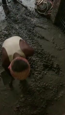 Three men are working with wet concrete on a construction site. One man is bent over and appears to be smoothing the concrete with his hands. Another man is using a shovel to move the concrete. A third man is standing and looking at the work. The ground is covered in wet concrete and mud. There are some bricks and construction materials visible in the background.