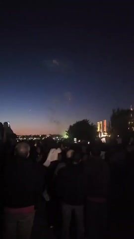 A nighttime scene shows a crowd of people watching fireworks. The sky is dark blue with hints of orange and pink near the horizon. Several bursts of colorful fireworks, primarily yellow, red, and green, illuminate the sky, leaving trails of smoke. In the foreground, the backs of several people are visible, some holding up cell phones to record the event. Trees and distant buildings with lights are silhouetted against the sky. The overall atmosphere appears festive, with people gathered to witness the fireworks display.