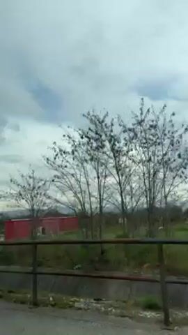 The video shows a view from a car's window. A yellow sign indicating "Golubovci" to the right and "Goricani" straight ahead is visible. The car seems to be driving along a road with a metal barrier and a drainage ditch beside it. In the background, there are some buildings, trees, and mountains under a cloudy sky. The camera then shifts to focus on the car's black leather headrest.