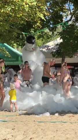 The video shows a group of people, including adults and children, enjoying a foam party on a sandy area, likely a beach or park. A large amount of white foam is being generated by a machine visible in the background, pouring down onto the participants. People are immersed in the foam, some appearing to be playing and interacting with each other. The environment includes trees providing shade and tents or structures in the background, suggesting an outdoor event. The overall atmosphere appears to be festive and recreational.