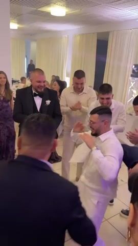 In a well-lit banquet room with a white ceiling, a group of at least nine people are dancing and celebrating. The room has white curtains and columns, with additional guests visible in the background. The lighting changes from green to purple to blue.

A man in a black suit and bow tie dances with a wide smile. A man in all white clothing dances energetically next to him, wearing glasses and a watch. Several other men in light-colored shirts clap and dance along. A drum set is visible to the left.

Two women in dresses stand near the dancers, and additional people are seen in the periphery. One man briefly ducks as part of the dancing. Overall, the atmosphere is festive and joyous.