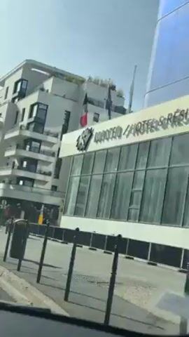 From inside a vehicle, a view of the exterior of the Kopster Hotel & Residence in Paris is visible. The building has white siding and a glass front. There are flags visible on the roof. A multi-story building can be seen next to the hotel.