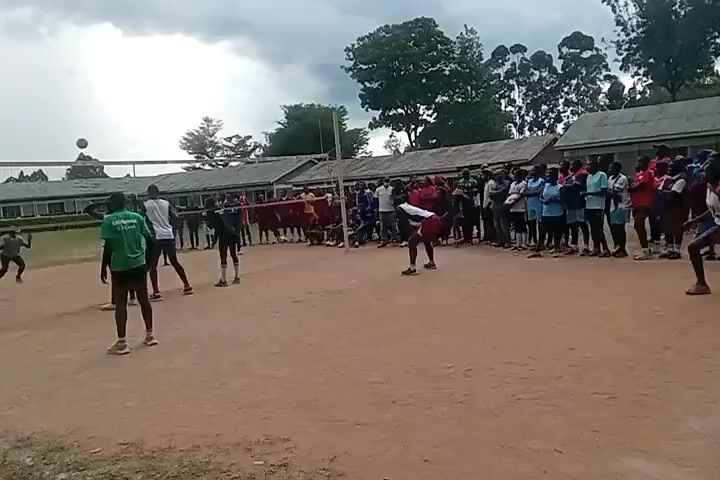 The video shows a volleyball game taking place on a dirt court. There are two teams of young people competing, with a net dividing the court. One team wears green shirts, while the other wears white and blue. A crowd of onlookers is gathered along the sidelines, observing the game. The sky is overcast, and there are buildings and trees in the background. One player spikes the ball over the net, and the crowd cheers.