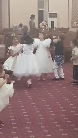 A group of young children, mostly girls in white dresses and one boy in a black shirt and patterned pants, are dancing on a red carpet with a patterned design. The girls are holding hands and spinning, while the boy is also dancing. Another young child in a white dress is standing to the left, facing away from the camera. In the background, there are chairs, a table with a floral arrangement, and some adult figures. The lighting appears to be artificial, and the overall environment seems to be an indoor event space, possibly a hall or ballroom.