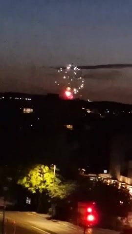 The video shows a nighttime scene with fireworks exploding in the distance over a dark, silhouetted landscape. In the foreground, there is a brightly lit tree on the left and a traffic light with its red light illuminated on the right. The sky is a deep blue transitioning to black, with a few faint clouds. The fireworks are various shades of white, yellow, and red, bursting in a clustered pattern against the dark sky. The overall impression is of a celebratory event taking place in a town or city.