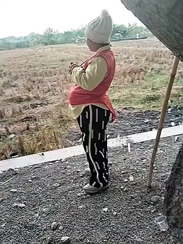 The video begins with a child wearing a light-colored beanie, a yellow shirt, a red vest, and black and white striped pants. The child is standing in an outdoor setting, facing away from the camera, with their hands clasped in front of them. The background shows a field with dry vegetation and some trees. The scene then transitions to a street with several people walking. A large green building is visible on the right side of the frame. Several banners are displayed, one of which appears to be for "Pondok Pesantren Nurul Huda." People of various ages, including children and adults, are seen walking along the street. Some individuals are interacting with each other, and there is a general sense of activity. The environment appears to be a public area, possibly near a school or community center.