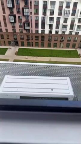A tabby cat stands on a balcony's railing, looking down at a playground. The cat then turns its head to look at the camera, extending a paw. The balcony is next to an outdoor air conditioning unit, with a modern apartment building in the background.