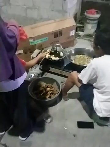 The video opens on a person wearing a purple headscarf and shirt, partially obscuring them as they stand near a gas stove. To their right, a child in a white t-shirt and dark shorts is seated on the ground, facing away from the camera, and looking at a wok filled with food being cooked. Another person, also wearing a purple headscarf and red shirt, is visible in the foreground on the right, facing away from the camera. In front of the person at the stove, there is a black stovetop with two burners. On the left burner, a pan contains white food items. On the right burner, a wok contains cooking food. To the left of the stove, a large metal pot contains fried food. The background shows a concrete wall, a blue plastic container, and cardboard boxes.

The scene then shifts to a wider outdoor area at night. A large blue tarp is spread on the ground. Several people are seated on the tarp, some of whom appear to be eating. A young child is visible running in the background. There are several food containers and a large water jug on the tarp. The area appears to be paved with concrete.