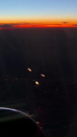 The video shows a view from an airplane window during sunset. The horizon is a vibrant band of orange and red, transitioning to a deep blue sky above. Below, the land is dark, with scattered city lights visible as small yellow dots. In the foreground, a portion of the airplane's wing or fuselage is visible, out of focus. Three blurry, bright orbs of light are also present in the middle ground, likely reflections on the window.