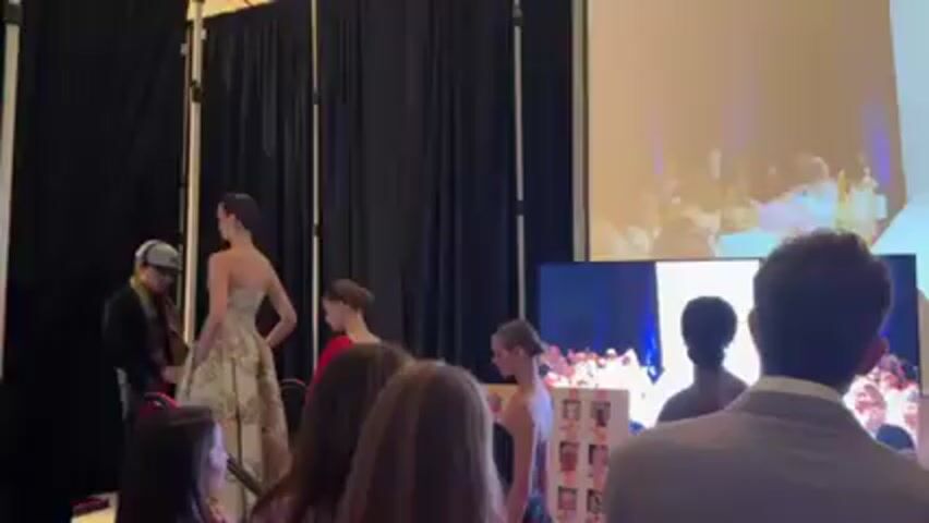 The video shows a group of people, mostly young women, standing in what appears to be a backstage area of a fashion show. Some of the women are dressed in formal wear, possibly as models. A man in a dark jacket is adjusting the dress of one of the models. Behind them, there are black curtains and some equipment like light stands. In the background, there is a large screen displaying a live feed of the fashion show, showing a model walking the runway. A smaller monitor also displays the show. The people in the video are watching the screen and seem to be either preparing for their turn or observing the ongoing show. There is also a board with images on it, likely related to the show's design or models. The room seems well-lit and professionally set up, suggesting a coordinated event.