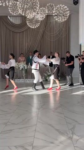 The video shows a wedding reception with a dance performance. Two couples are dancing a traditional folk dance in the foreground, dressed in embroidered vests and skirts. In the background, a wedding party is seated at a decorated table, and two videographers are filming the performance. The room is elegantly decorated with draped curtains and elaborate light fixtures.