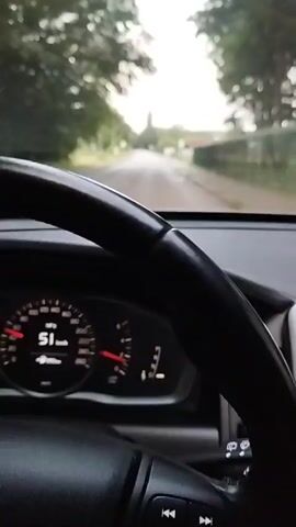 The video shows a first-person perspective from inside a car. The dashboard displays the speedometer at 50 km/h. The car is driving on a paved road lined with trees on the left and a fence and buildings on the right. The sky is overcast. In the distance, a tall structure resembling a tower or steeple is visible. The video captures the road ahead and the interior of the car, including the steering wheel and parts of the dashboard. The scene appears to be a suburban or rural area.